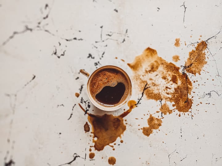 A top-down photo of a cup of coffee and coffee stains on a white, scratched up surface. 