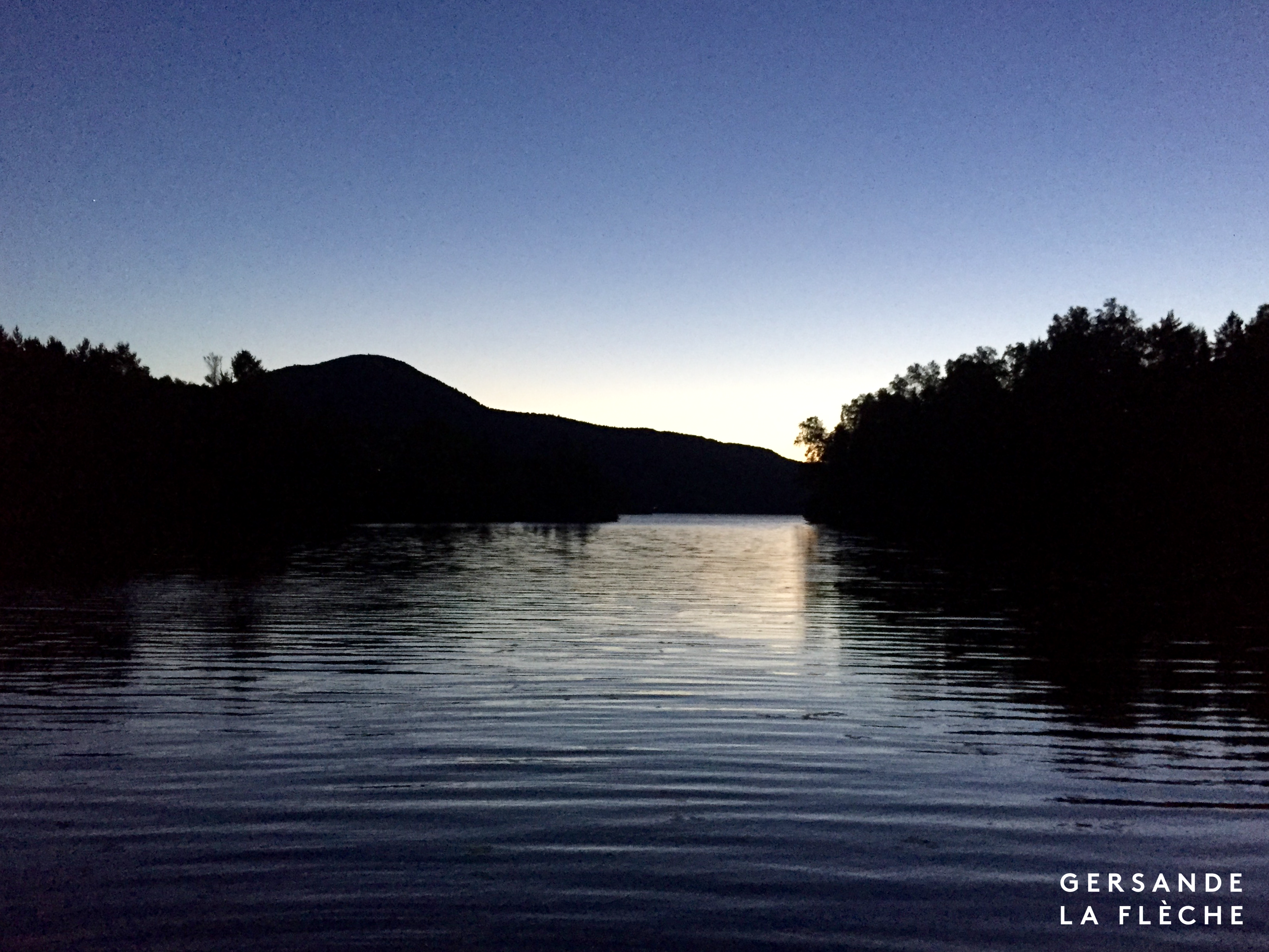 A photo of a mountain and a lake after sunset, with the silhouette of the forest reflected on the still silver-bright surface of the water.