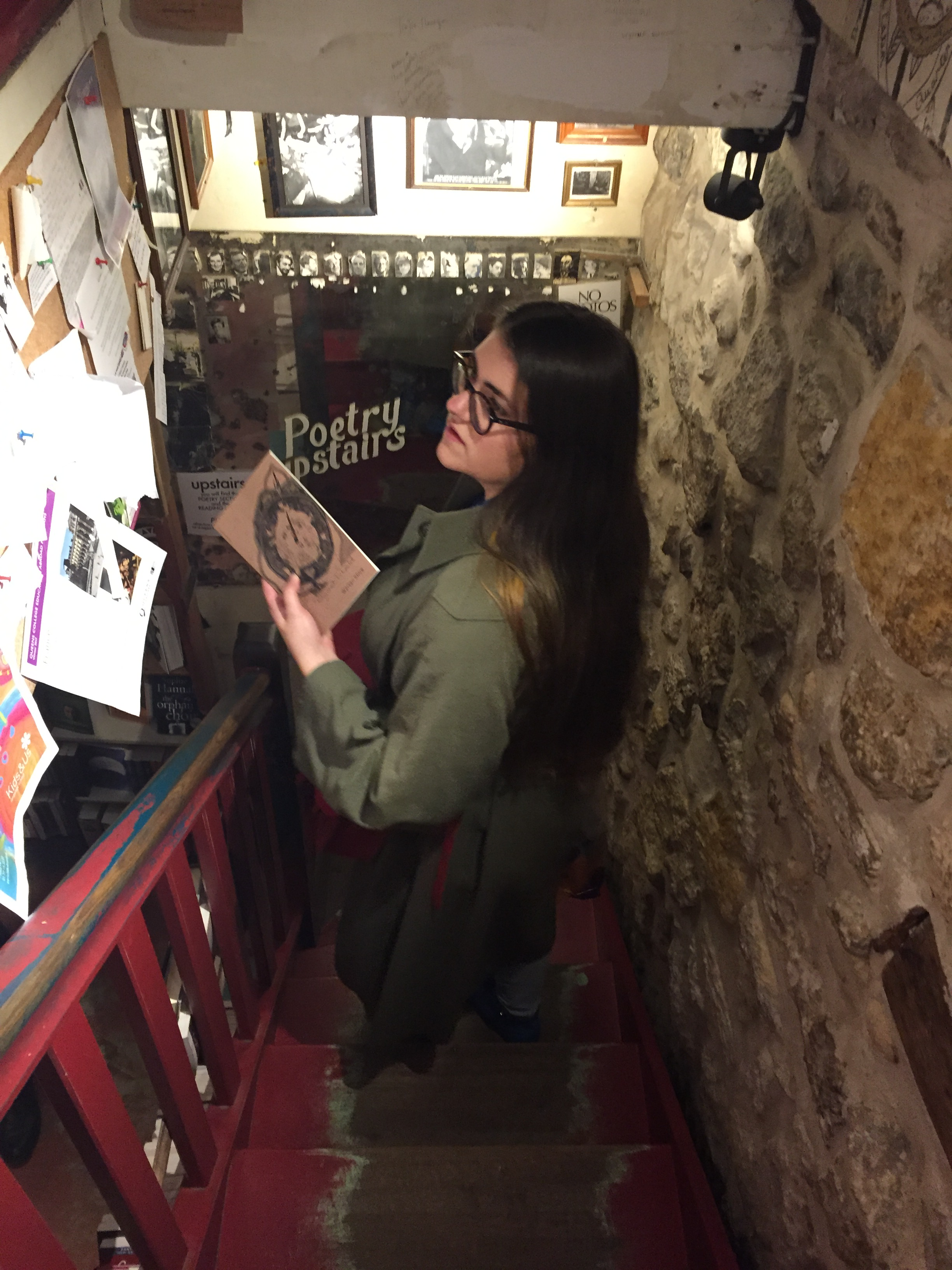 A picture of me on a staircase in Shakespeare and Co, holding a copy of Warsan Shire's "teaching my mother how to give birth" and reading the bookstore's community board.