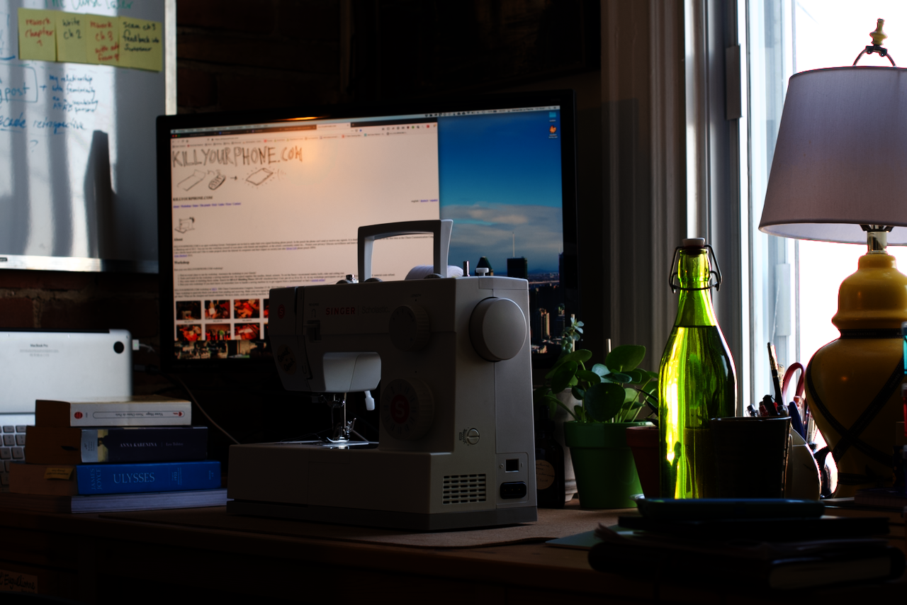 A photo of my desk, with my little Singer Scholastic sitting upon it. There are a bunch of books and notebooks on the desk, a bottle of water, several plants, and on the computer screen behind and just above the sewing machine is the website KillYourPhone.com just barely out of focus. 