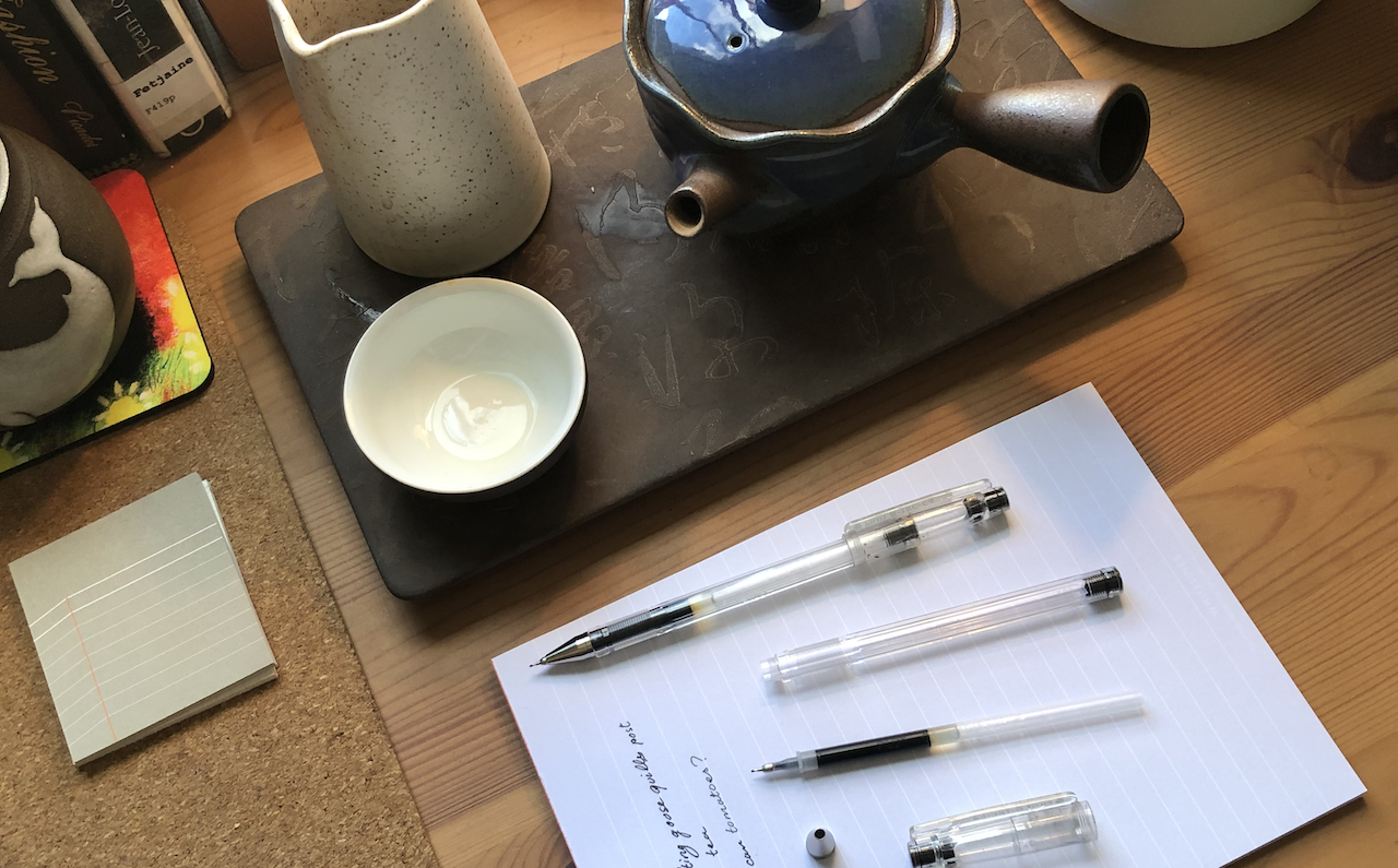 A photo of my desk table, showing some tea cups, a teapot, paper and sticky notes, as well as two Pilot G-Tec pens, one disassembled.