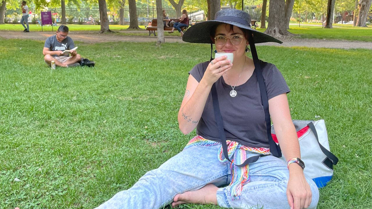 A photo of me smiling at the camera, sitting down on the grass, wearing a big black sunhat and drinking tea out of a little ceramic mug in my hand.