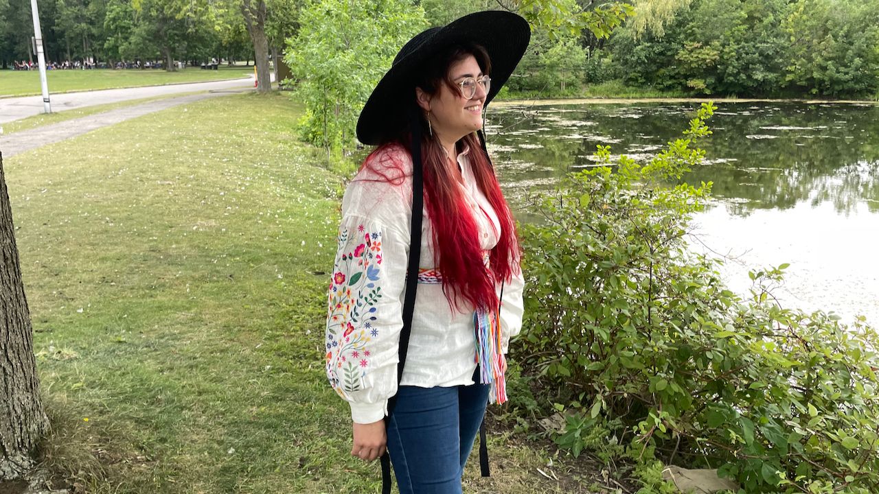 A photo of me standing in a park by a pond, wearing a black sunhat and white shirt with brightly coloured sleeves.