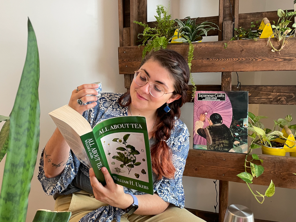 Myself, Gersande, with hair in a side braid and wearing a blue patterned top, reading a book surrounded by plants and tea.