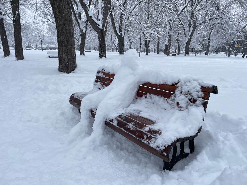 On a park bench, a sculpture of a snowman is chilling happily and watching the snow fall.
