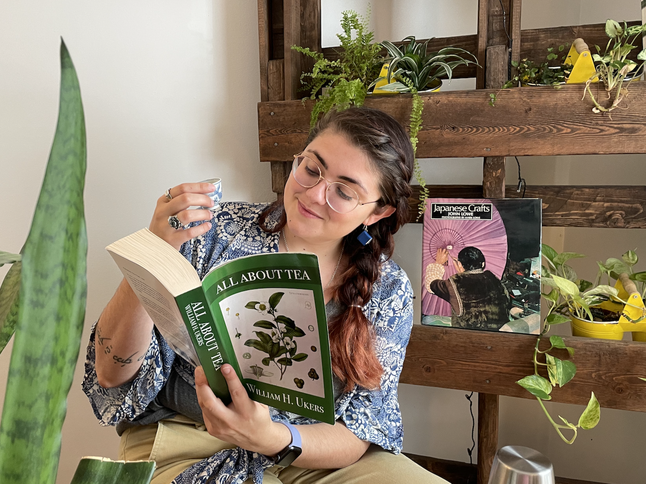 A photo of me sitting and reading a book, tiny tea cup in hand, surrounded by many green plants. I'm wearing little book-shapped earrings and the book is called All About Tea.