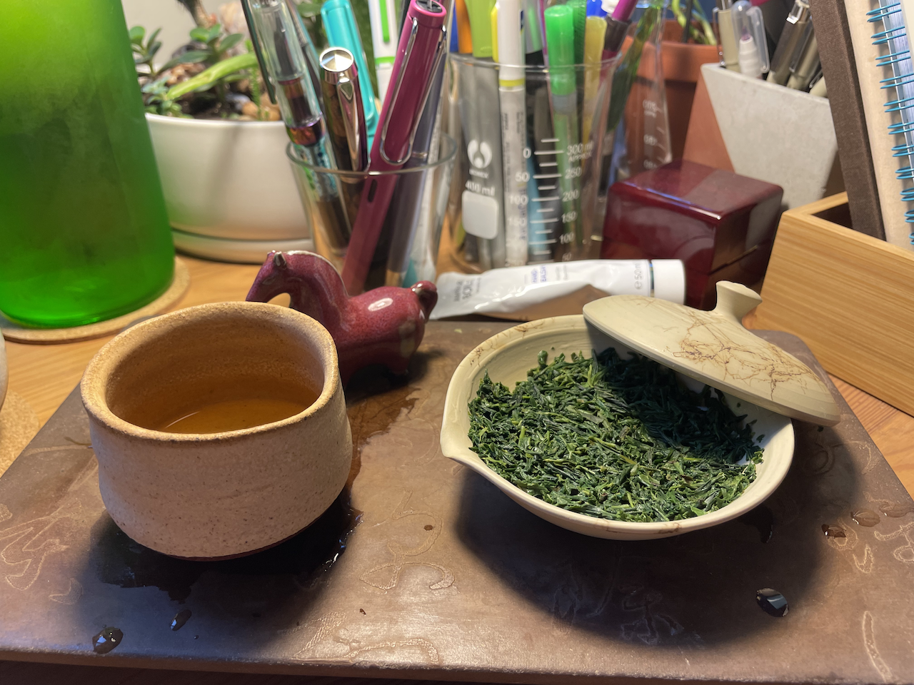 A photo of my tea tray, featuring a little ceramic horse tea pet, a little ceramic cup in an earthy beige colour and of rough texture, and a white and beige speckled shiboridashi that can hold a little under 100mL of water. In this picture, the cover of the shiboridashi is tilted and propped on the side of the bottom, so that you can see the deep green leaves of the gyokuro after the first infusion.